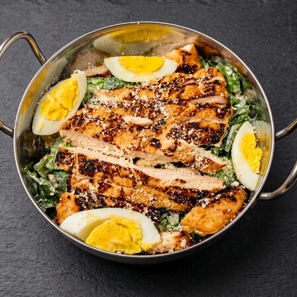 Grilled chicken Caesar salad with hard-boiled eggs and parmesan cheese in a metal bowl on a dark surface.