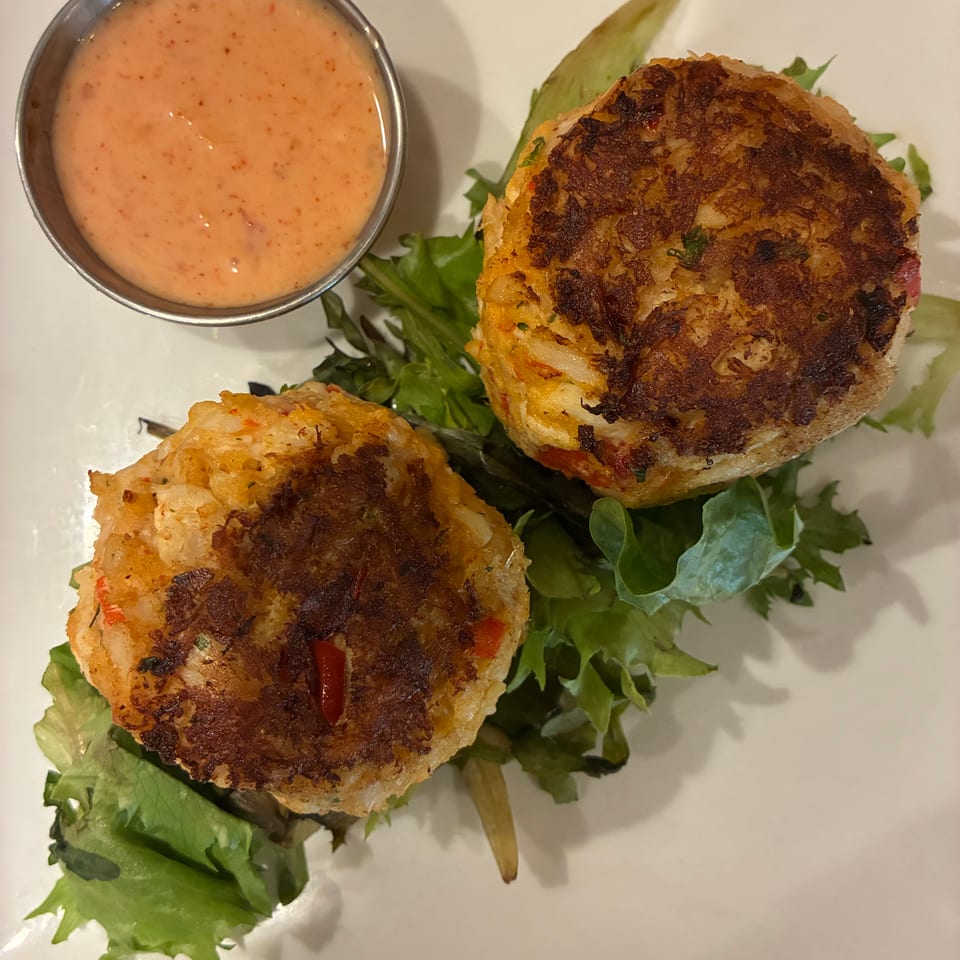 Two golden-brown crab cakes on a bed of green lettuce with a side of creamy orange sauce.