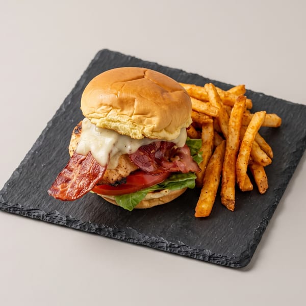 Grilled chicken burger with bacon, melted cheese, lettuce, tomato, and a side of seasoned fries on a slate.