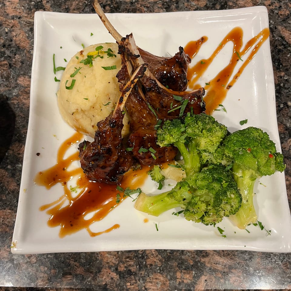 Glazed lamb chops with mashed potatoes and broccoli, drizzled with sauce on a white plate.